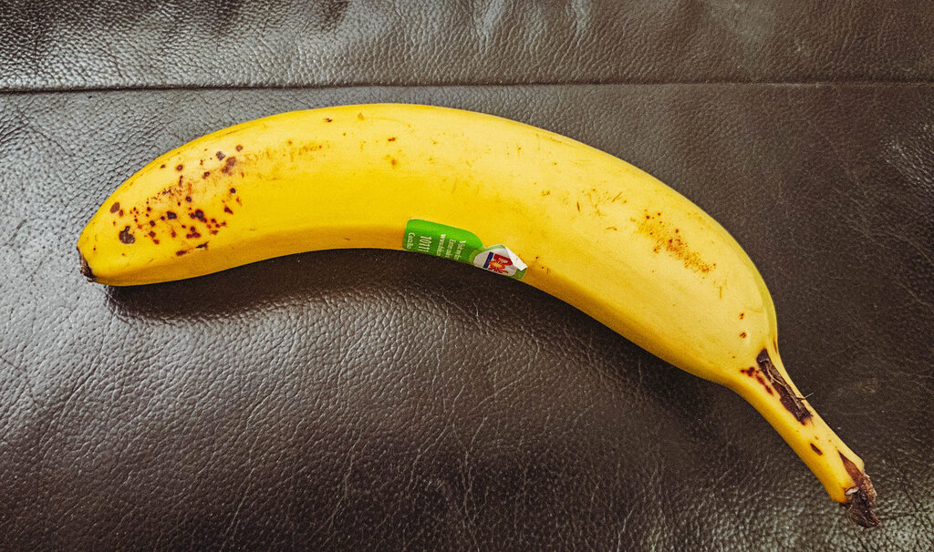 A single ripe yellow banana with brown spots resting on a dark textured surface, with a small green produce sticker attached
